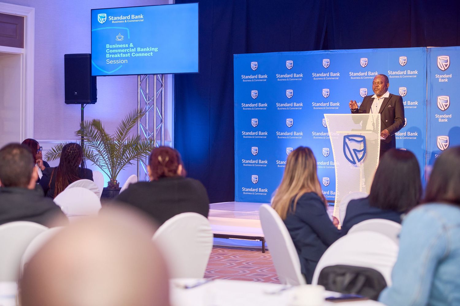 A speaker presents at a Standard Bank conference event to a seated audience in a formal banquet hall.