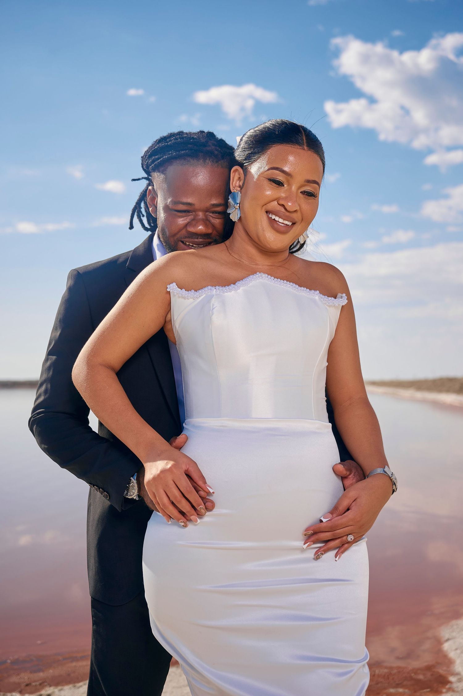Couple in wedding attire embracing by a pink lake under blue sky, bride in white strapless gown, groom in black suit.
