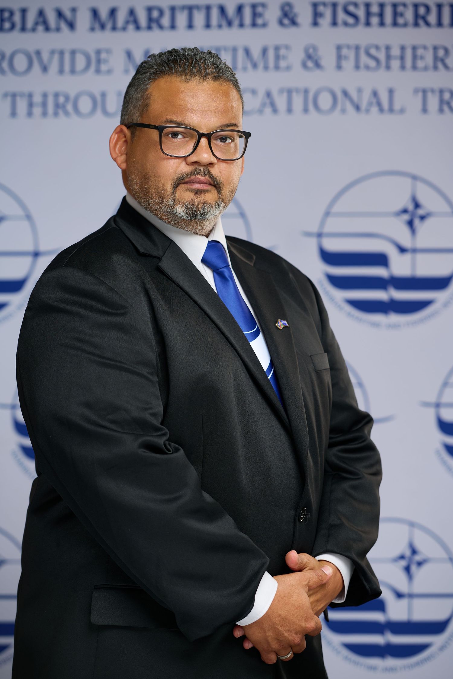 Professional wearing a dark suit and blue tie stands against a maritime-themed backdrop with company branding.