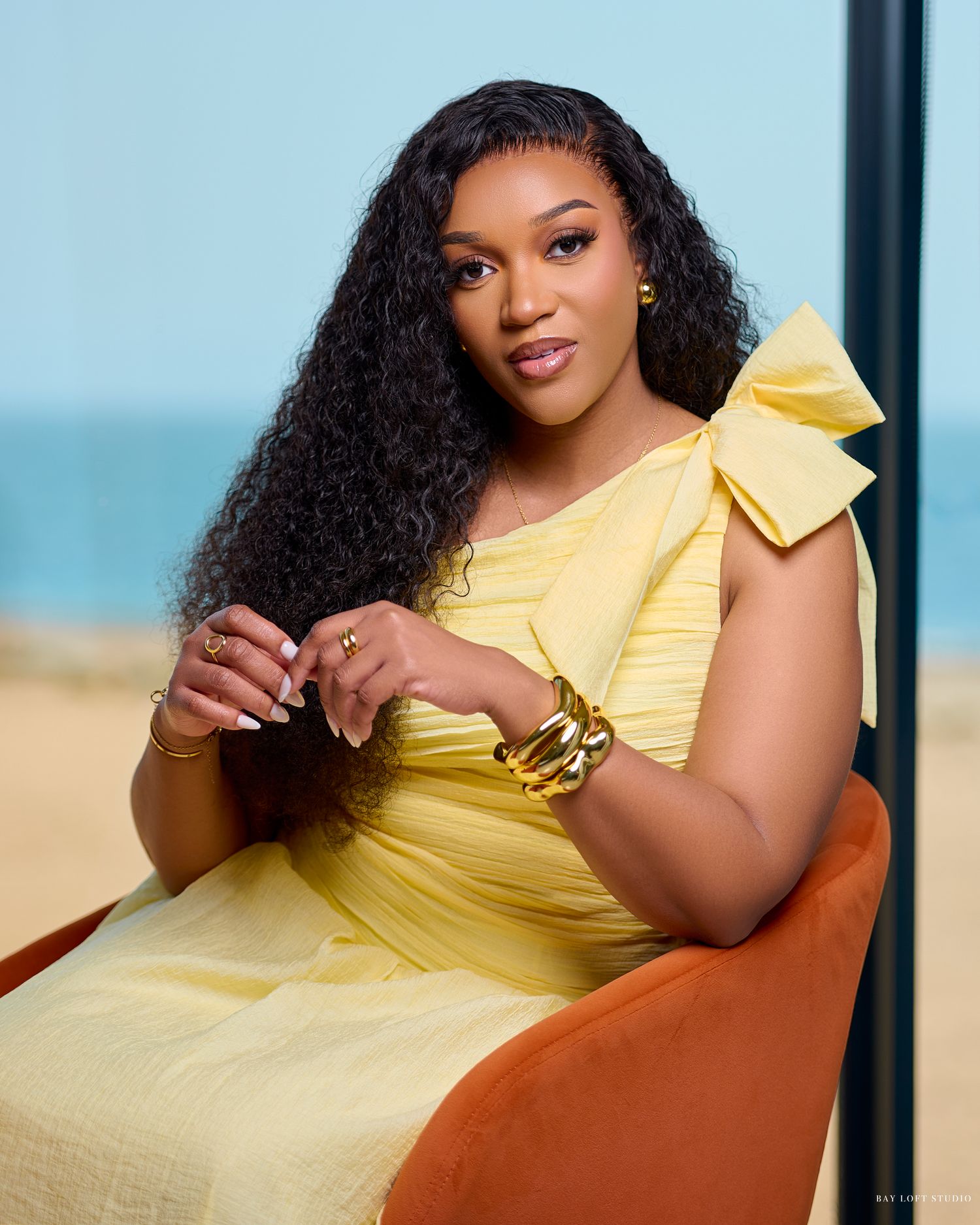 A person in a yellow dress with a bow detail sits on an orange chair against a beachy backdrop.
