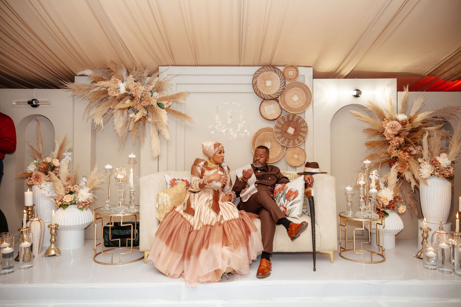Elegant couple in formal attire seated at a decorated wedding reception table with floral arrangements and gold accents.
