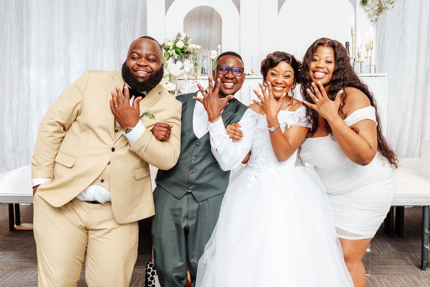 Joyful wedding party of four posing together, bride in white gown with guests celebrating at elegant venue.