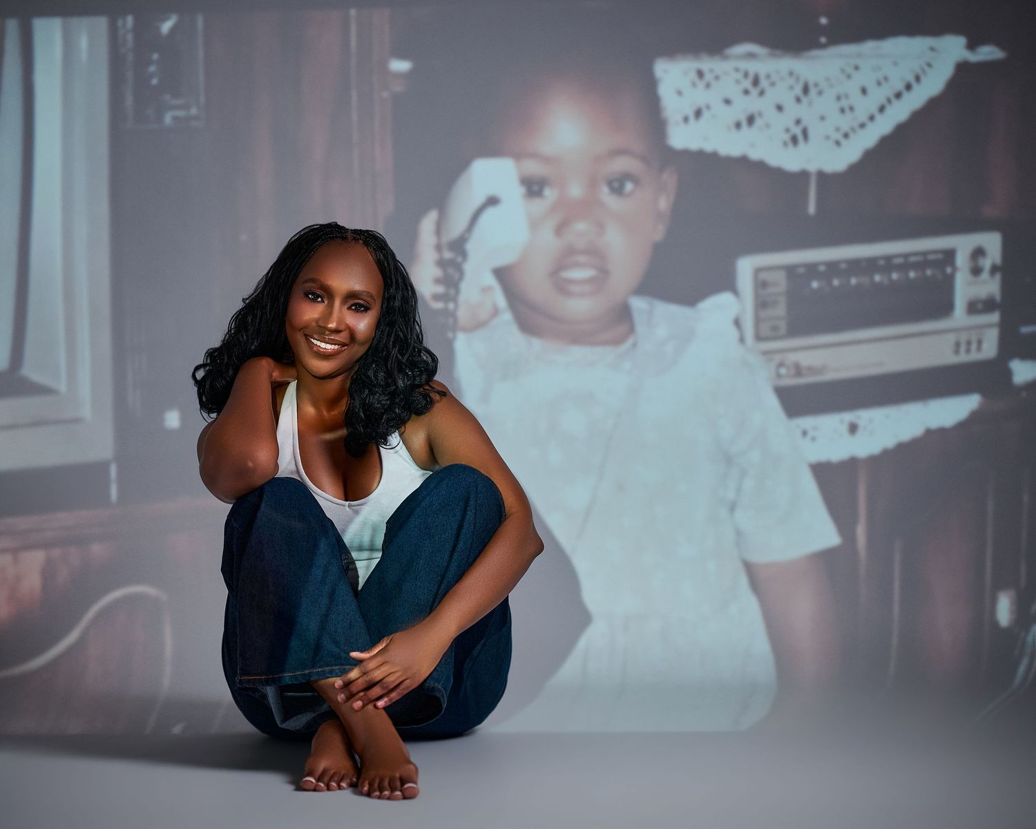 A woman sits cross-legged in front of a projected childhood photo of herself as a toddler holding a phone.