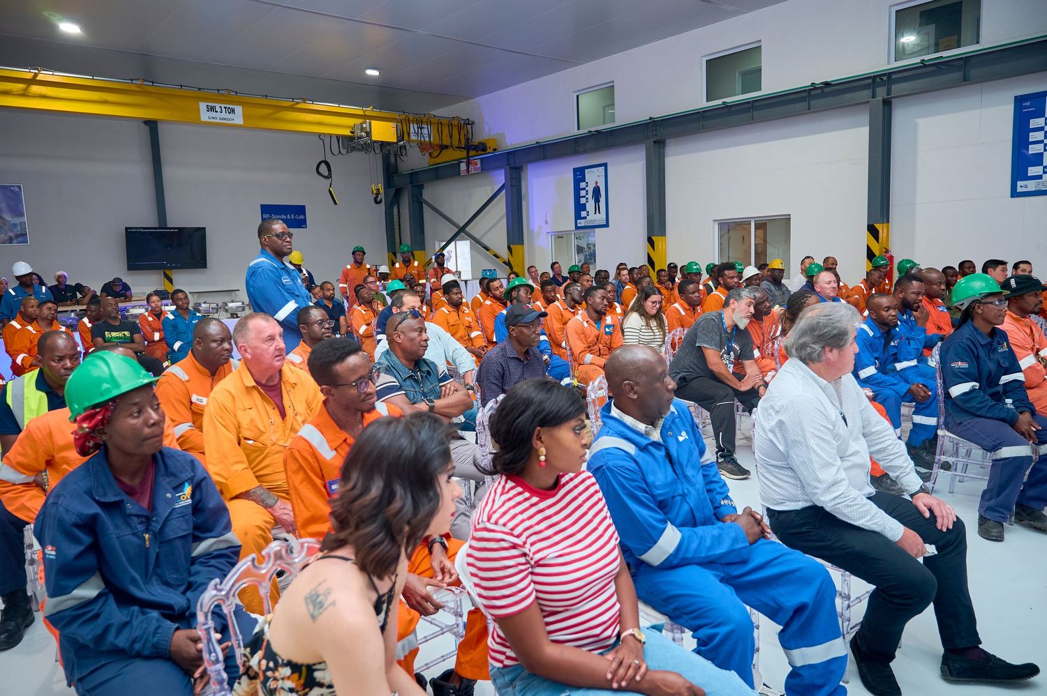 Large group of workers in orange uniforms attend a safety meeting inside an industrial warehouse.