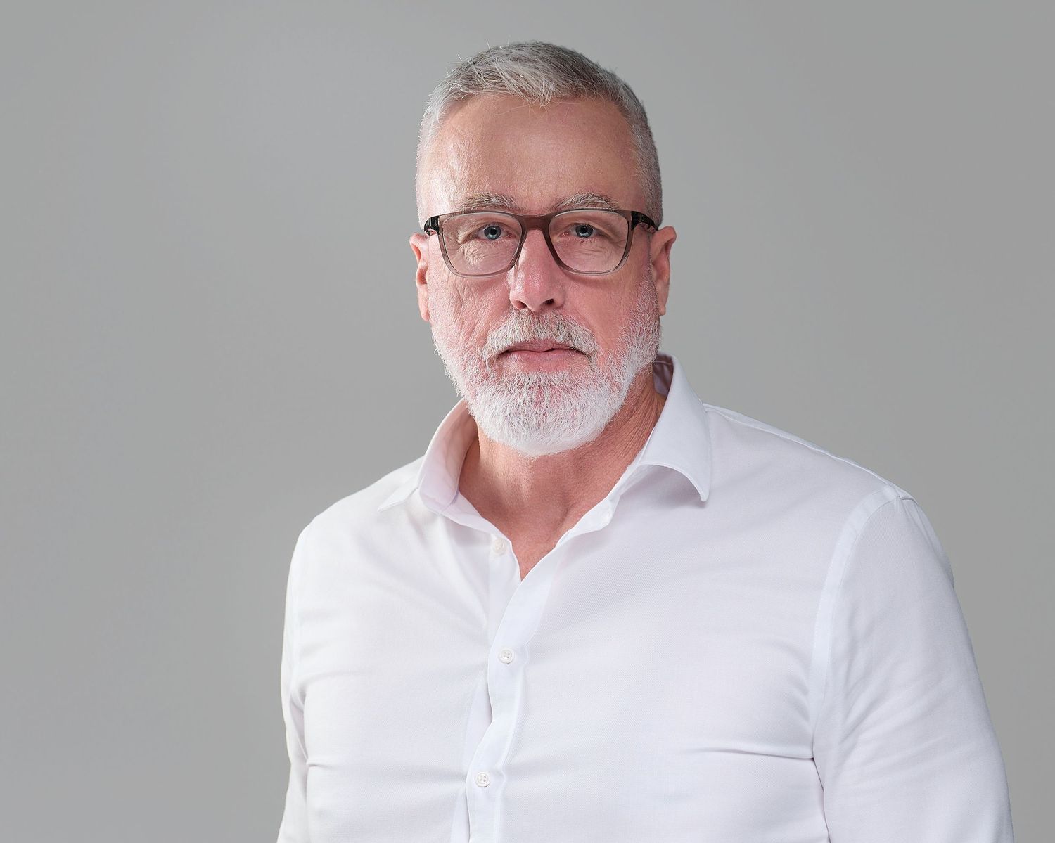Mature man with gray hair and beard wearing glasses and white shirt against gray background.