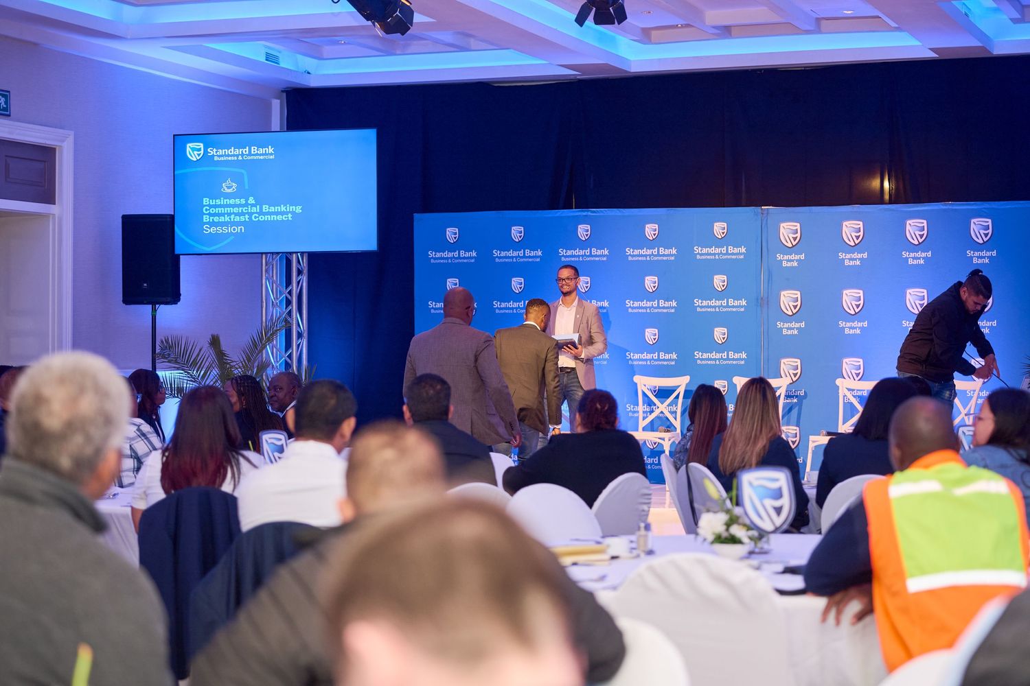 Attendees gather at a Standard Bank corporate conference with a presenter on stage in a decorated hall.