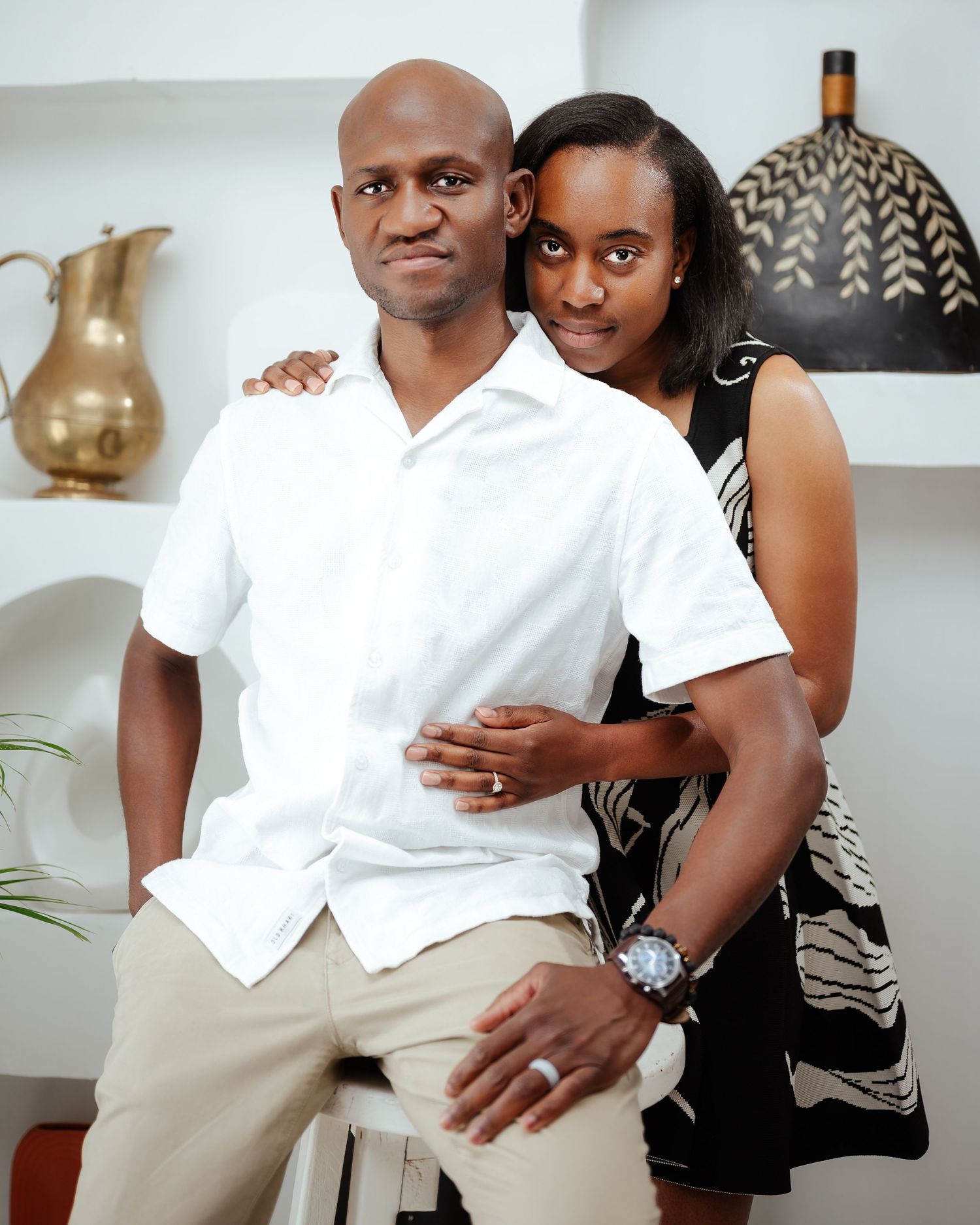 A couple poses together, man in white shirt and khaki pants, woman in black dress, in a bright modern interior.