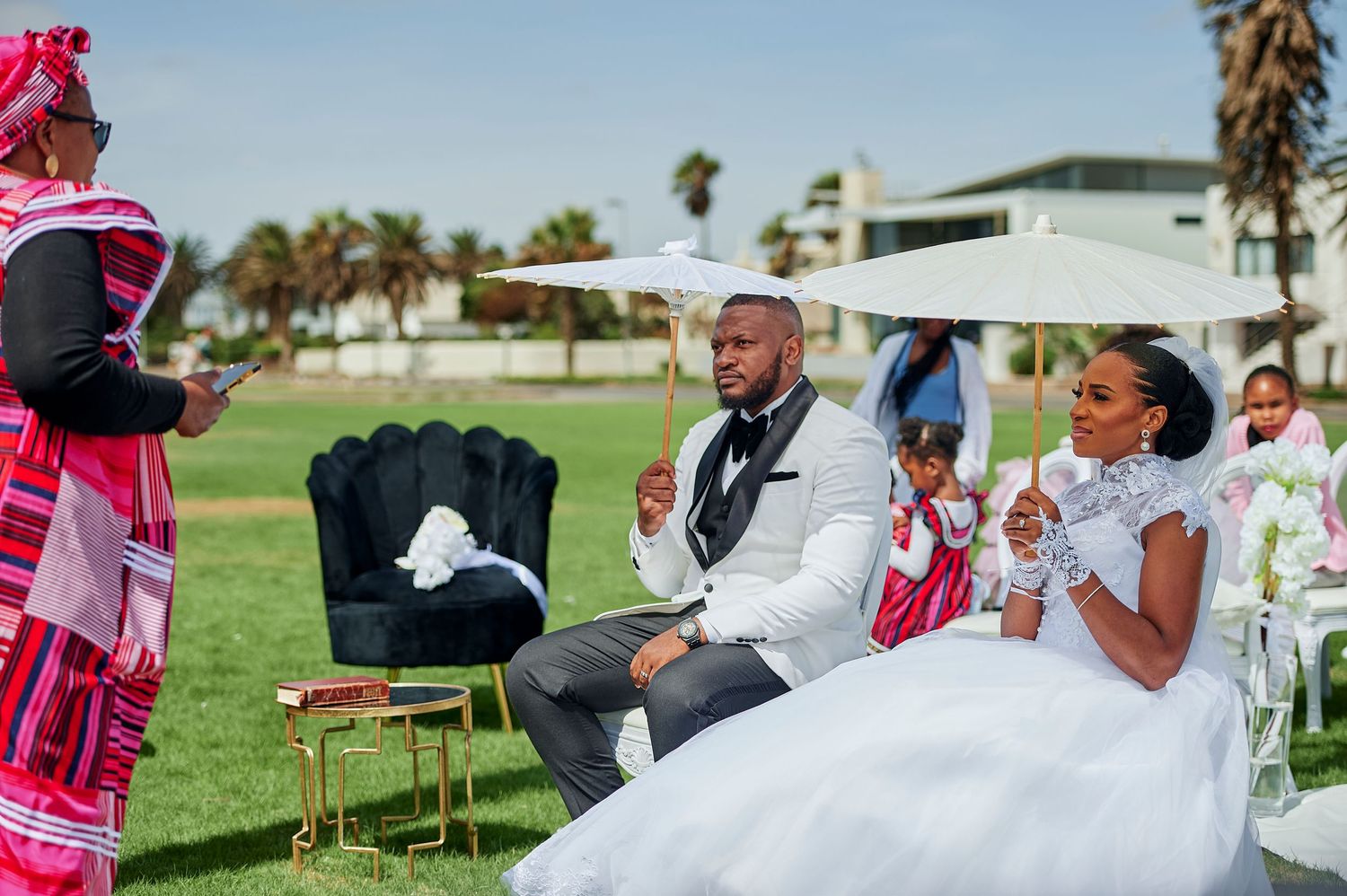 Bride and groom seated outdoors at a traditional African wedding ceremony with colorful attire and white umbrellas.