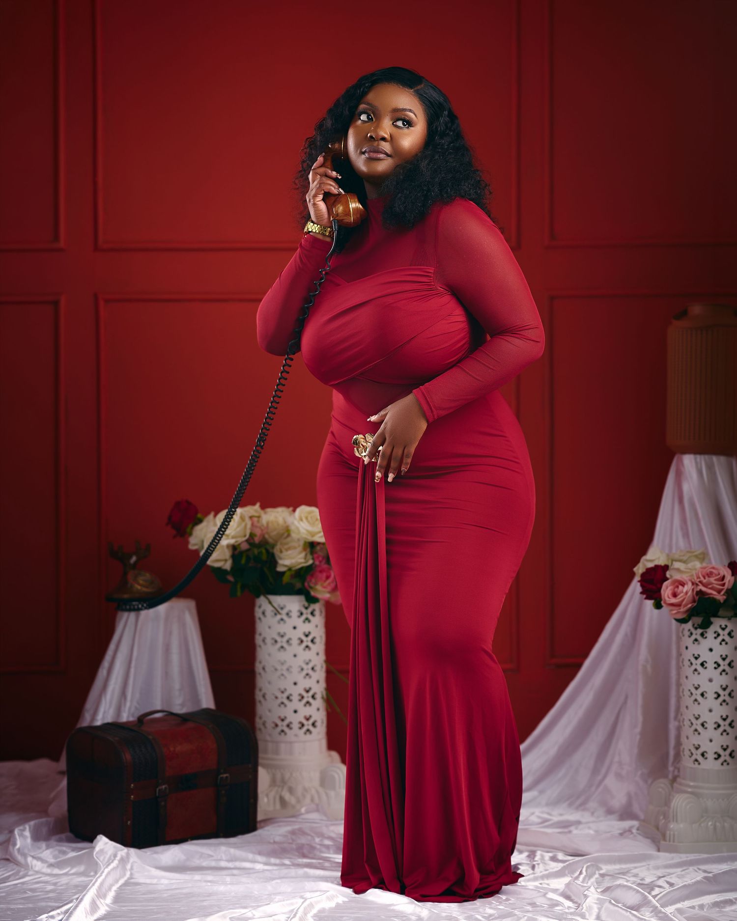 A plus-size woman in a red long-sleeve mermaid gown poses with a vintage phone against a deep red backdrop.