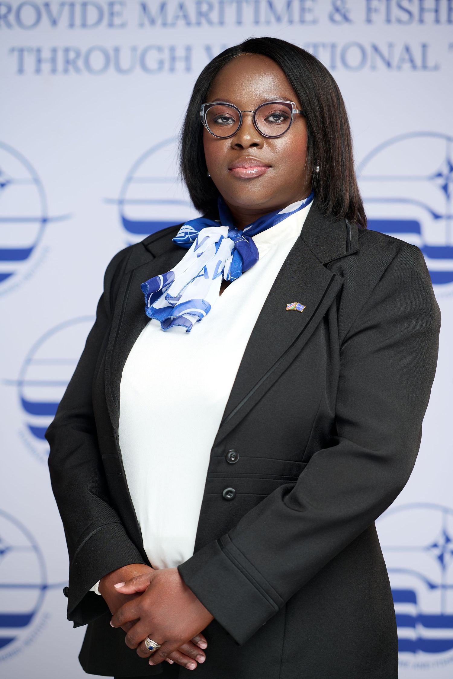 Professional woman in black blazer and blue scarf poses before a maritime and fisheries organization backdrop.