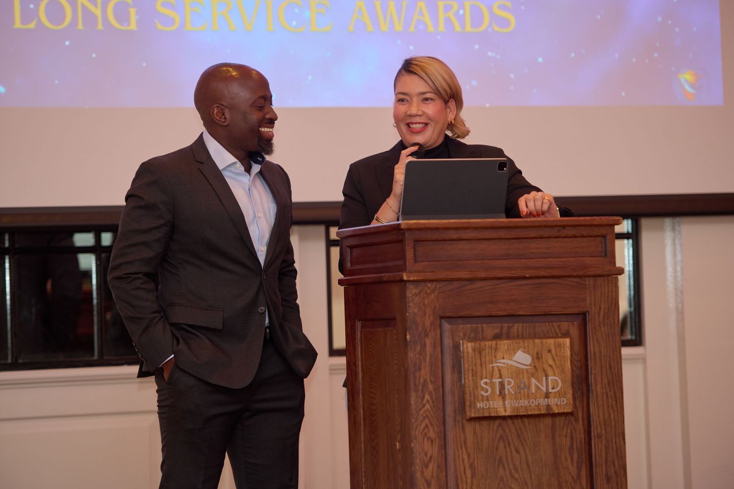 Two colleagues share a laugh at a Long Service Awards ceremony podium in a formal event setting.