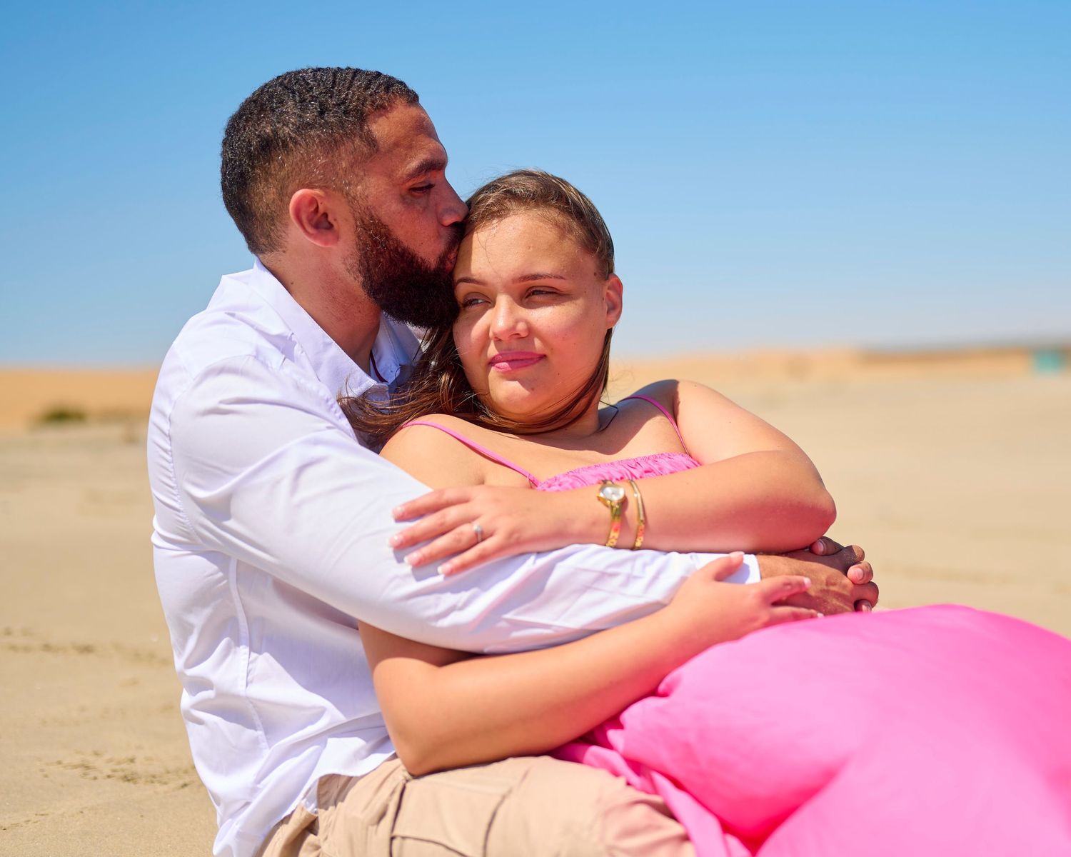 Couple embracing romantically on sandy desert dunes, man in white shirt kissing woman's cheek under clear blue sky.