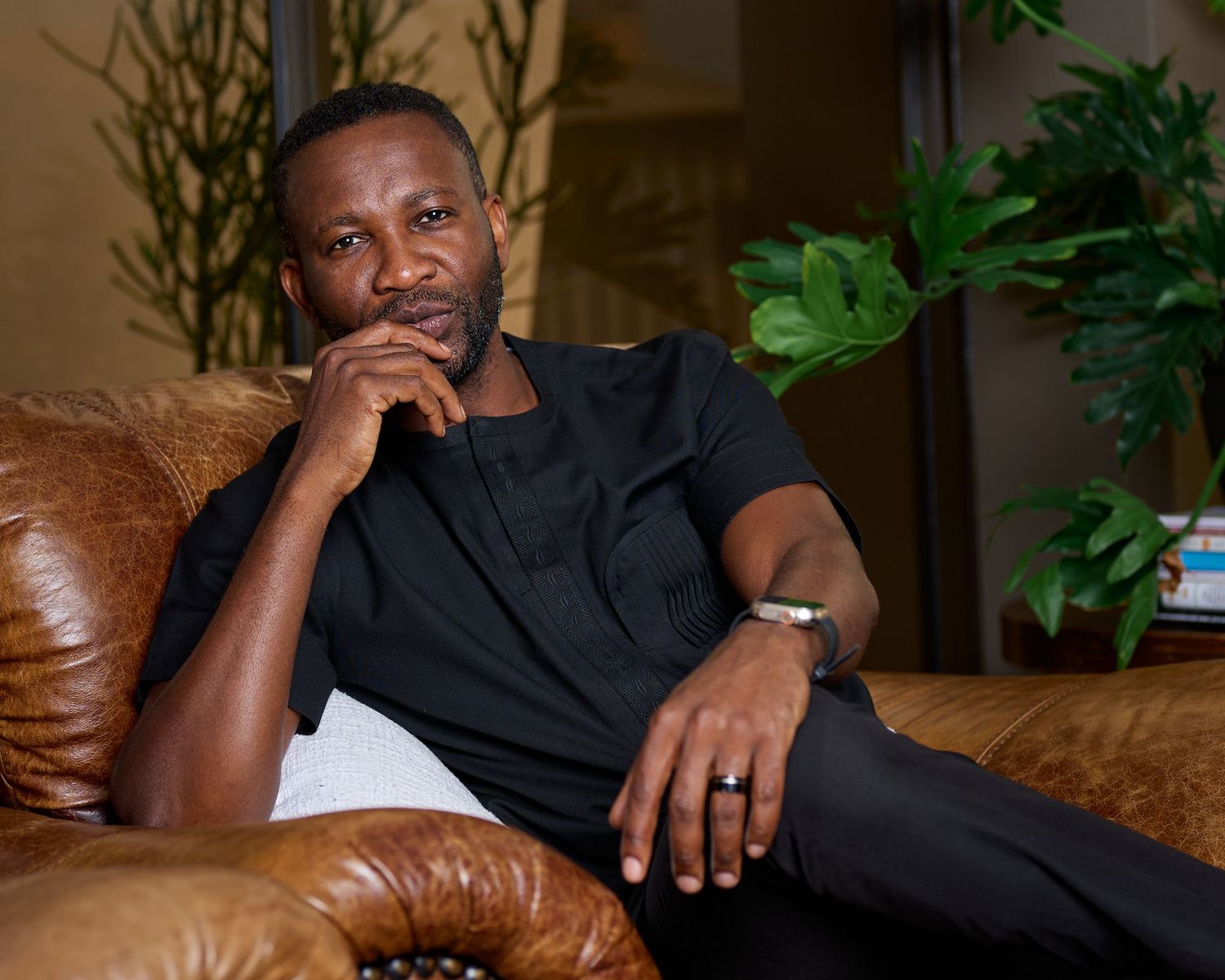A thoughtful man in a black shirt sits on a brown leather sofa, resting his chin on his hand.
