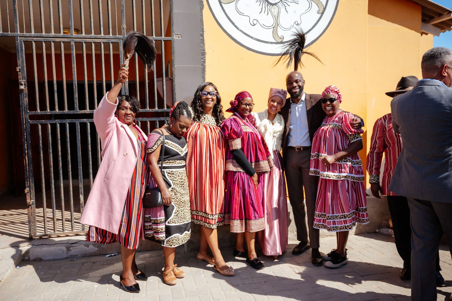 Group of people in vibrant traditional African clothing posing together outside a yellow building.