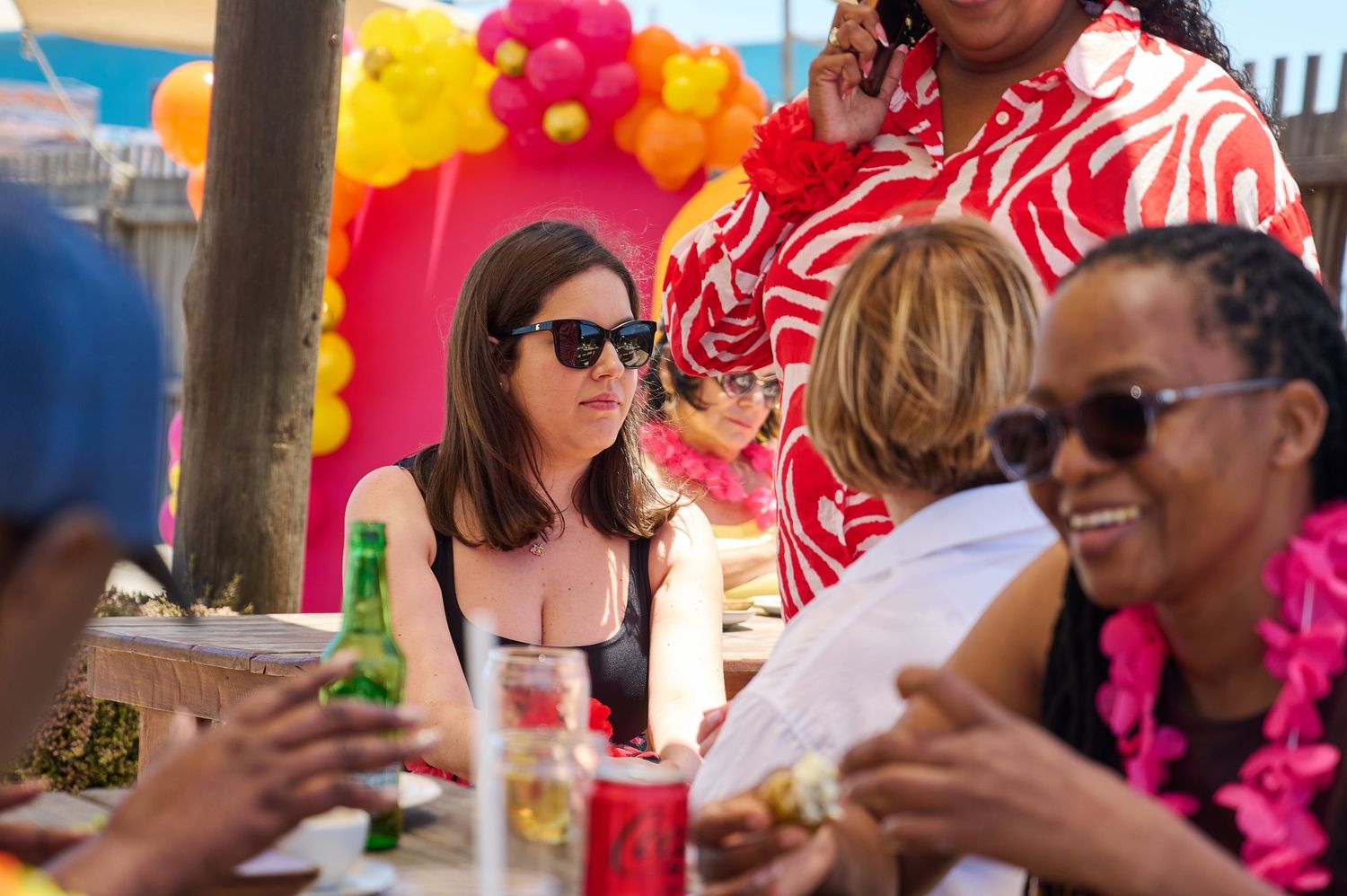 Diverse group of women socializing at a colorful outdoor party with balloon arch and tropical decor.
