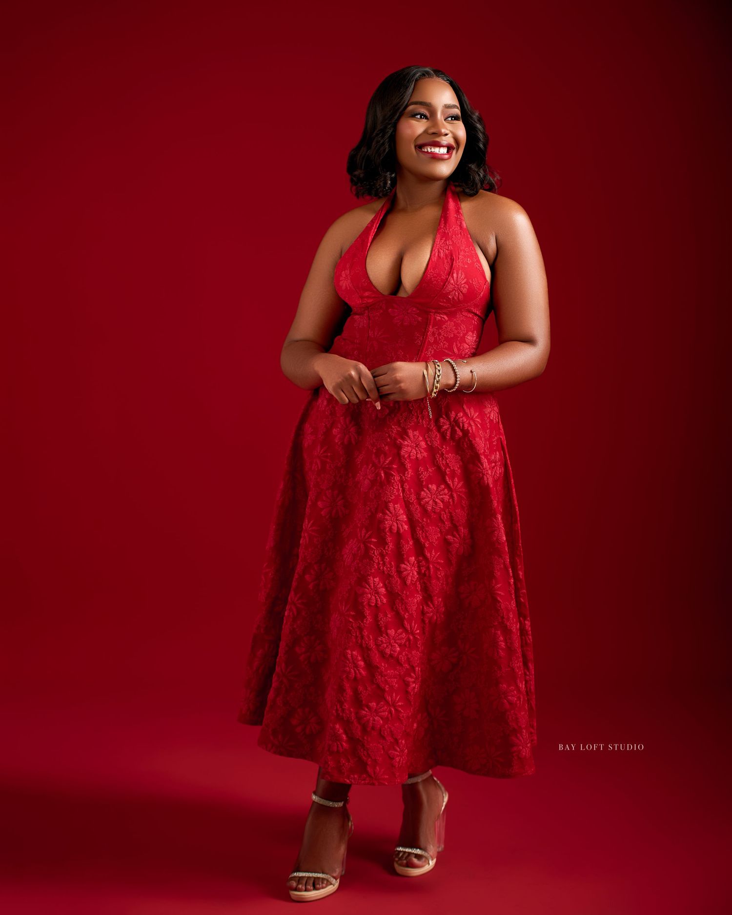 A woman in a red lace dress poses confidently against a deep burgundy background during a glamorous photoshoot.