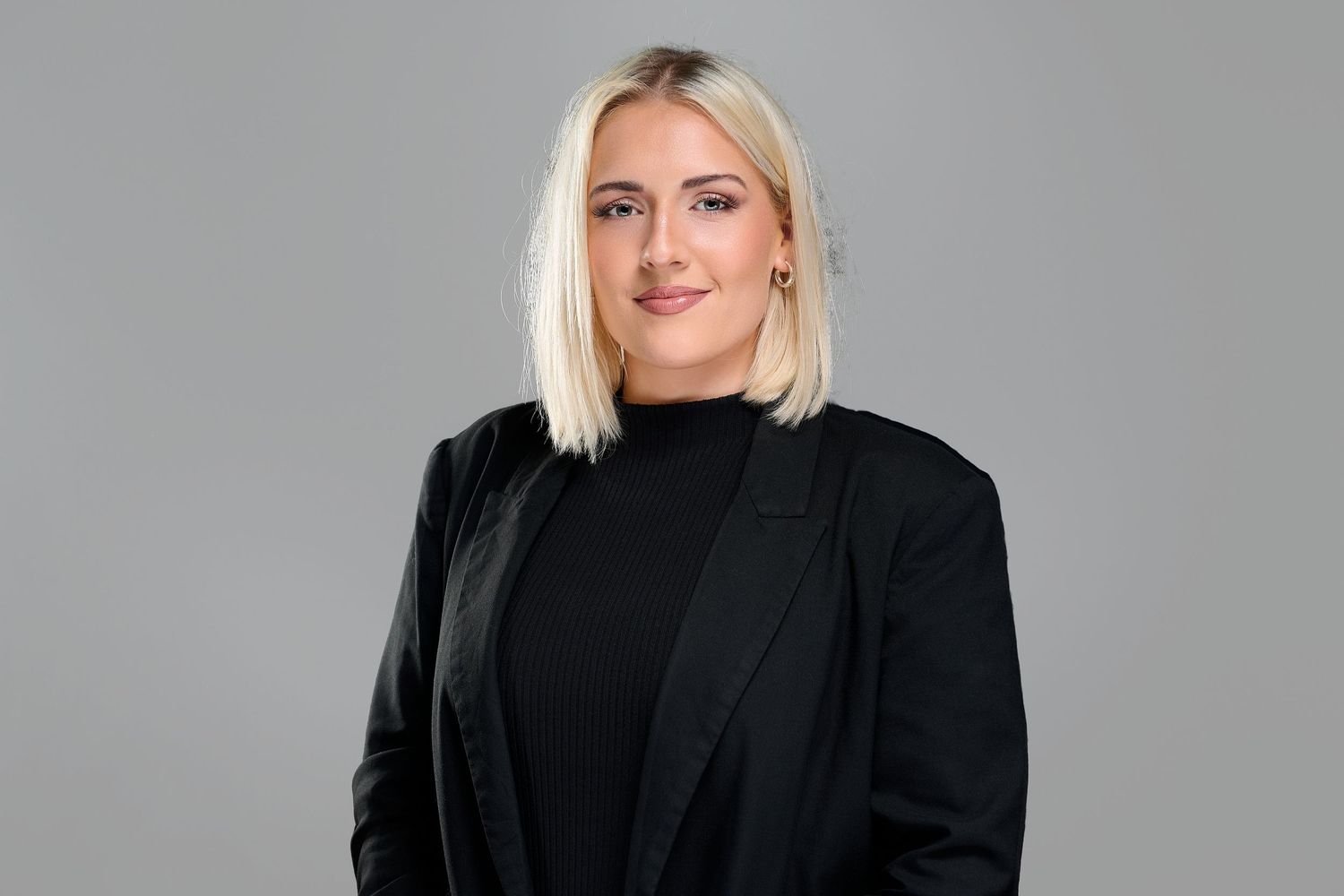 Professional woman with blonde bob haircut wearing black turtleneck and blazer against gray background.