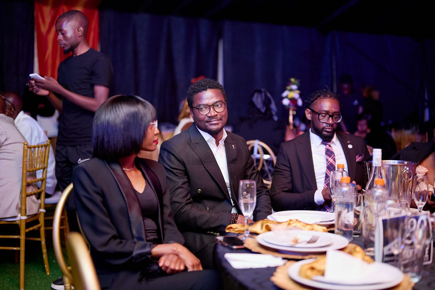 Three guests in formal wear sit together smiling at a decorated banquet dinner table.