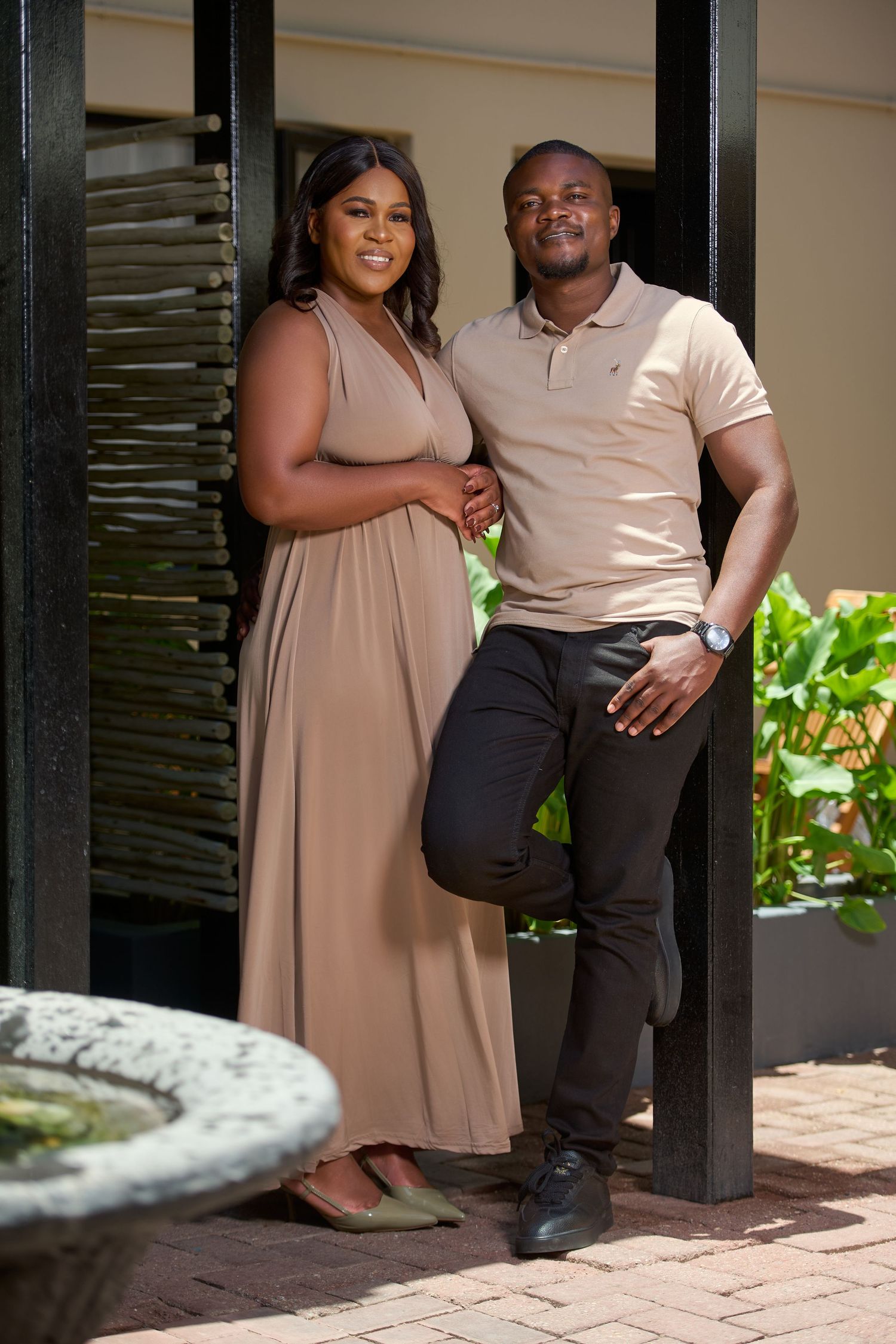 A smiling couple in matching beige outfits poses together by a dark pillar in an outdoor setting.