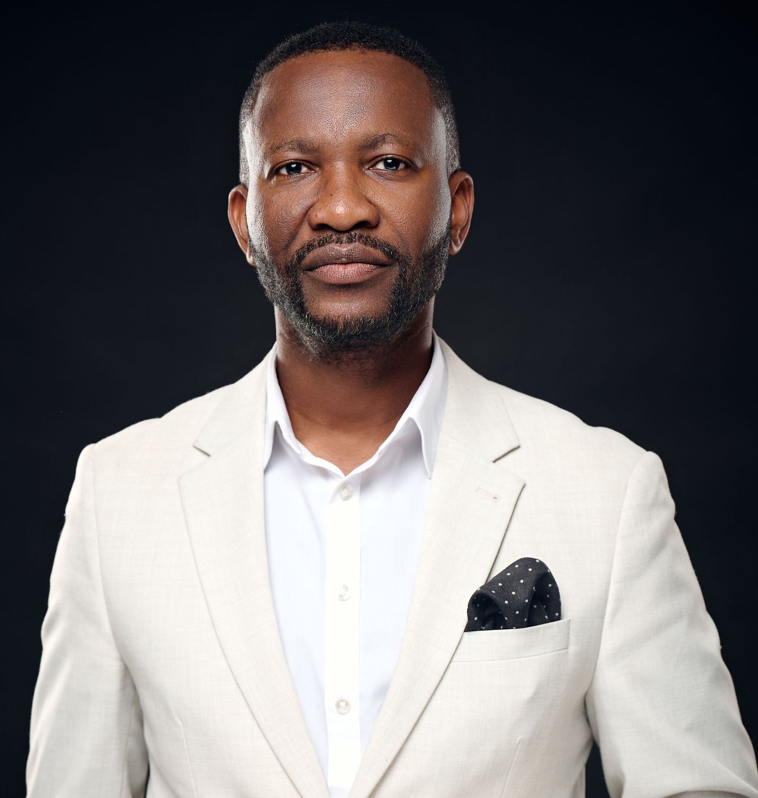 Professional man in cream blazer and white shirt poses confidently against dark background in formal portrait.