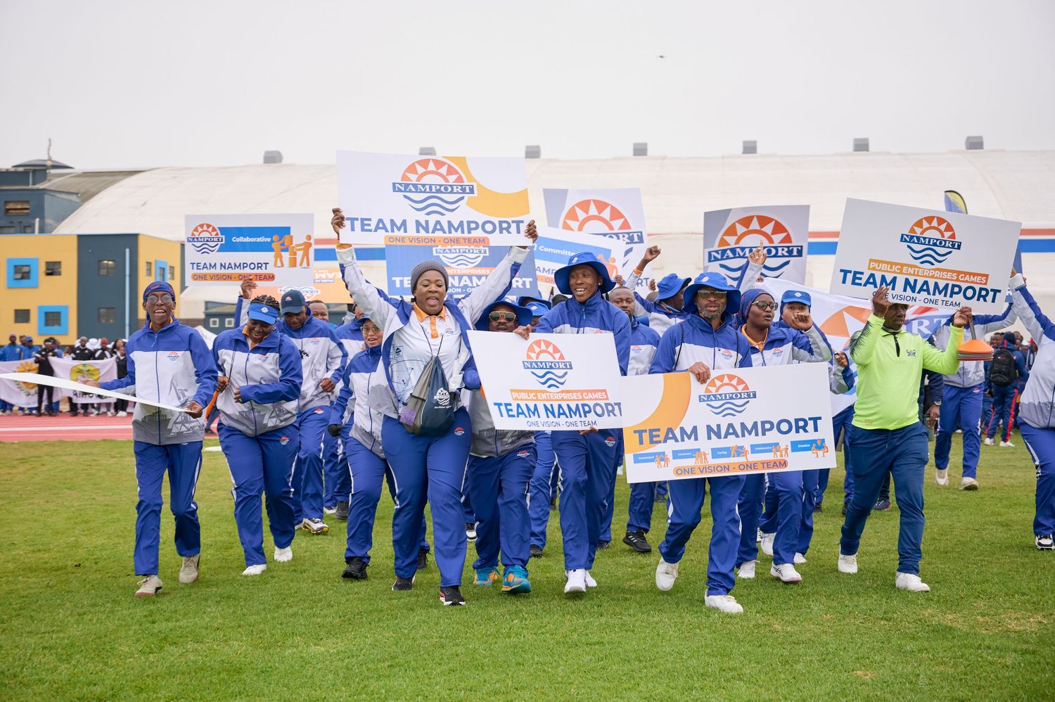 Team Namport members in blue uniforms proudly marching and holding banners at an outdoor sports event.