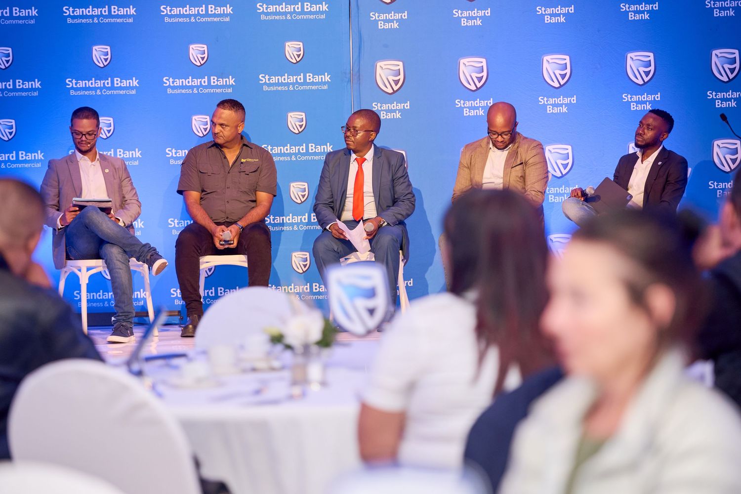 Five professionals sit in a panel discussion at a Standard Bank corporate event with branded backdrop.
