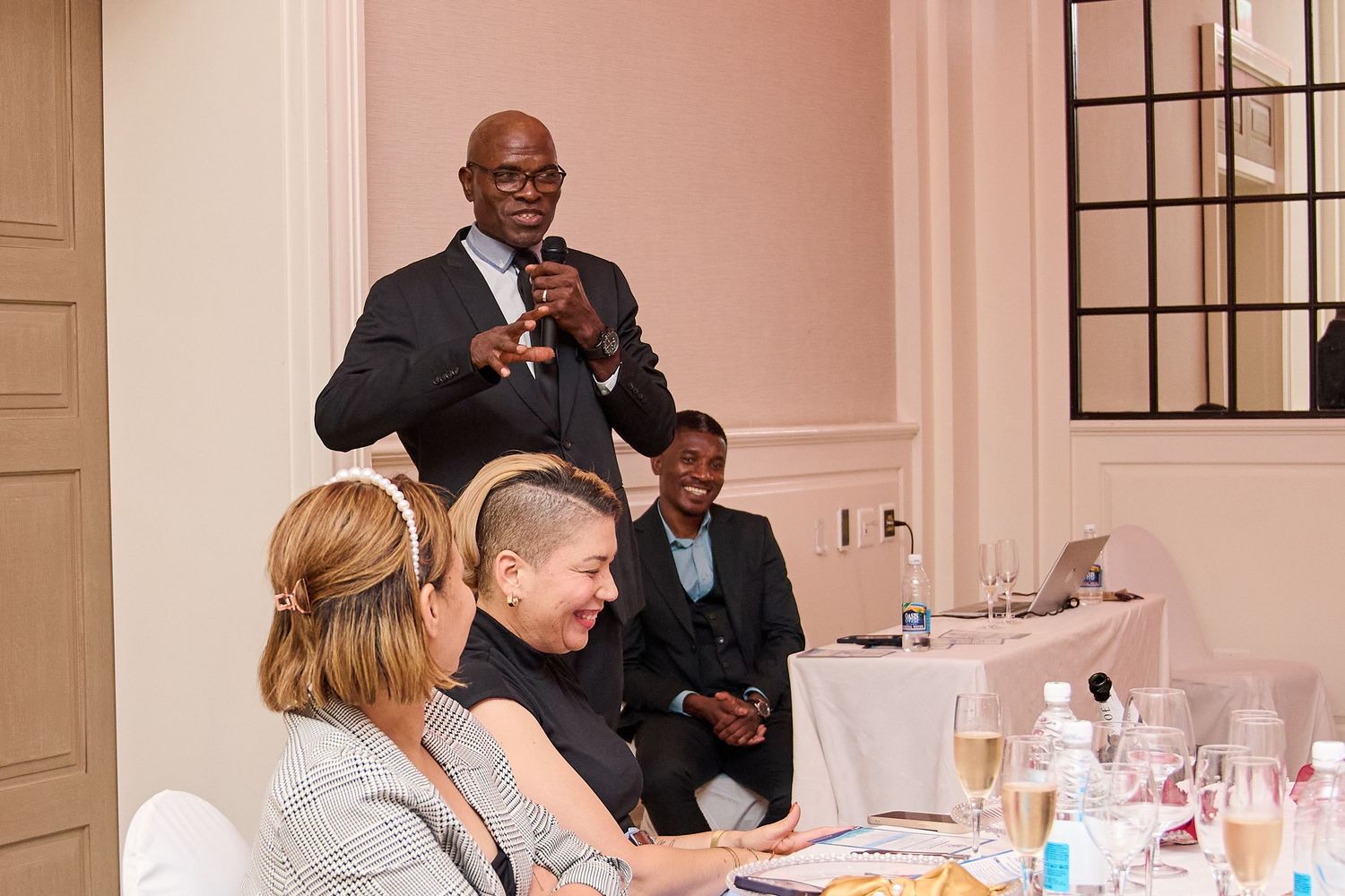 Man giving speech at formal dinner event, holding microphone while guests smile and laugh at decorated table.