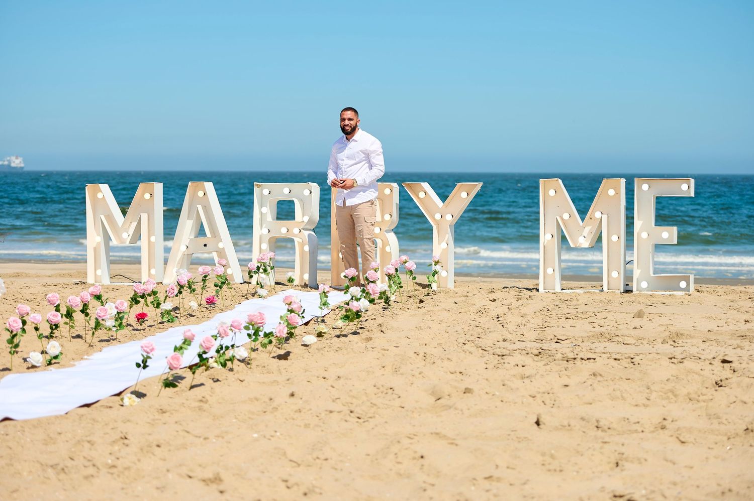 Man stands on beach beside large MARRY ME marquee letters decorated with pink flowers.