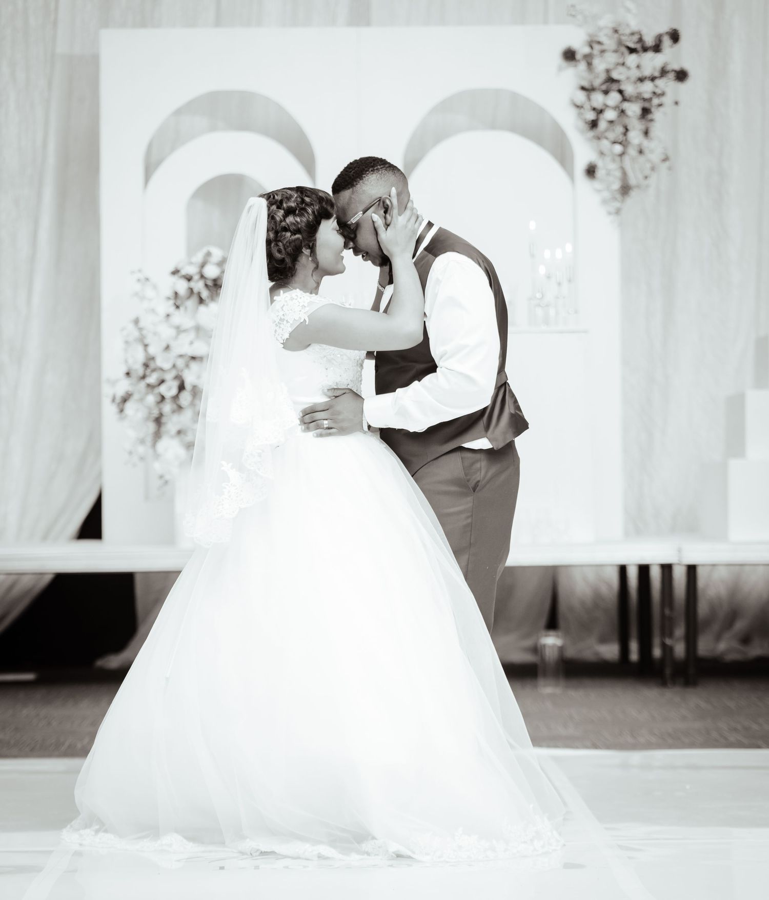 Black and white wedding photo of bride in white gown and groom in vest sharing a kiss at the altar.
