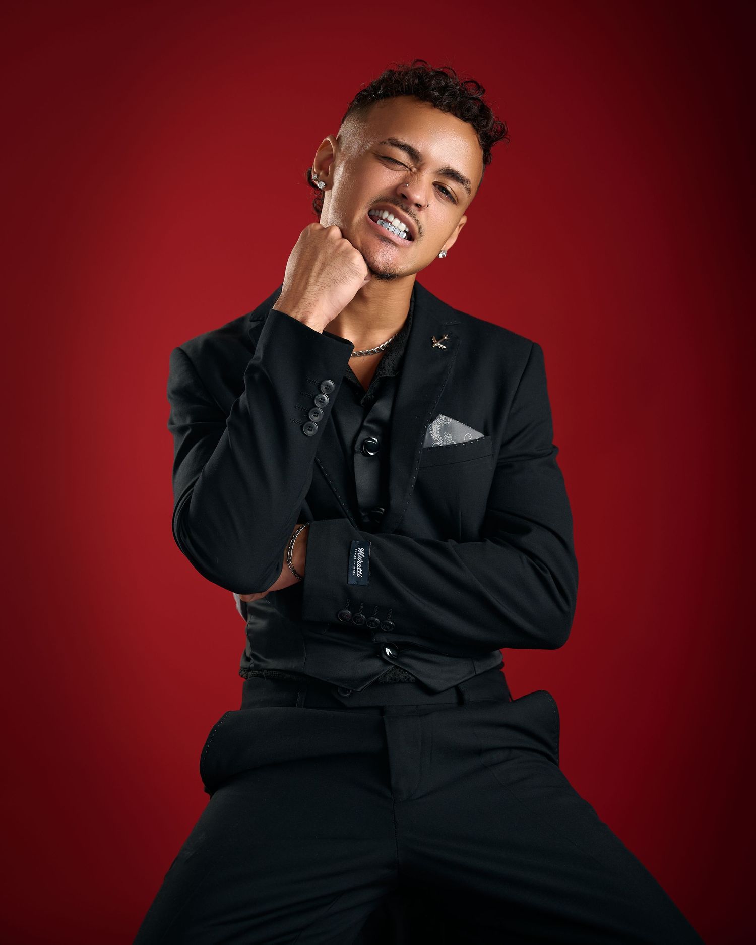 Young man in black suit with silver grillz posing confidently against a red background.