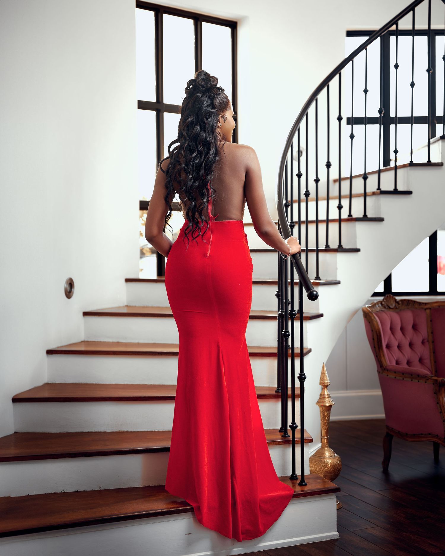 Woman in elegant red backless mermaid gown standing on staircase with curved iron railing.