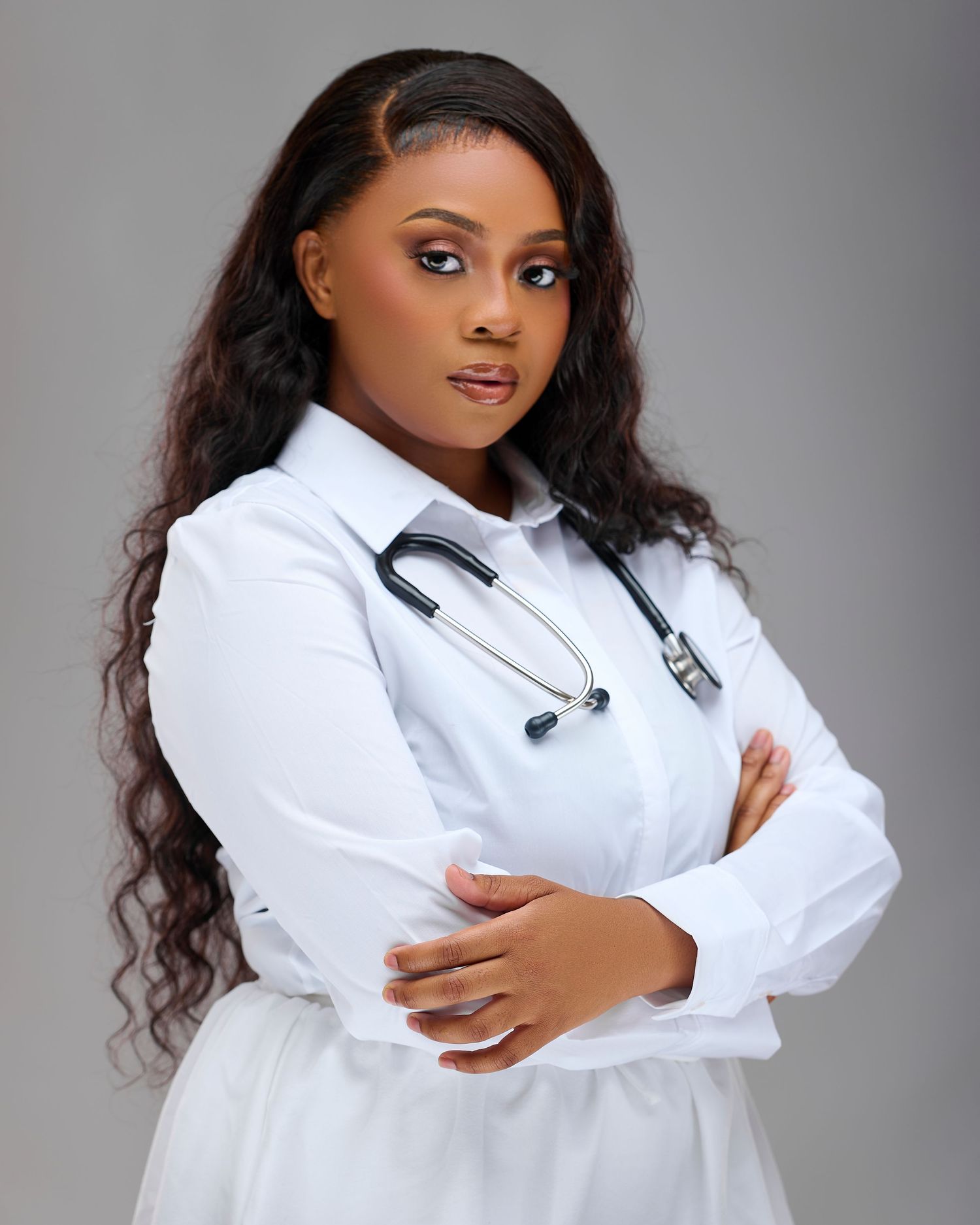 Young female doctor in white coat with stethoscope, arms crossed, posing confidently against gray background.