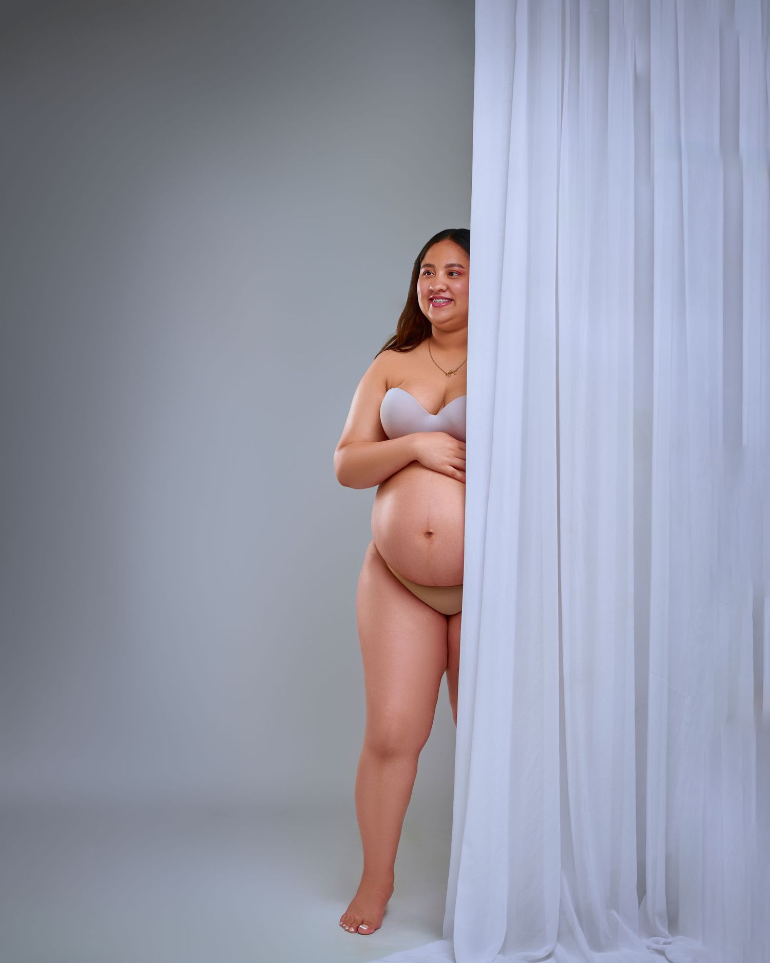 A person poses playfully behind a white sheer curtain in a studio setting.