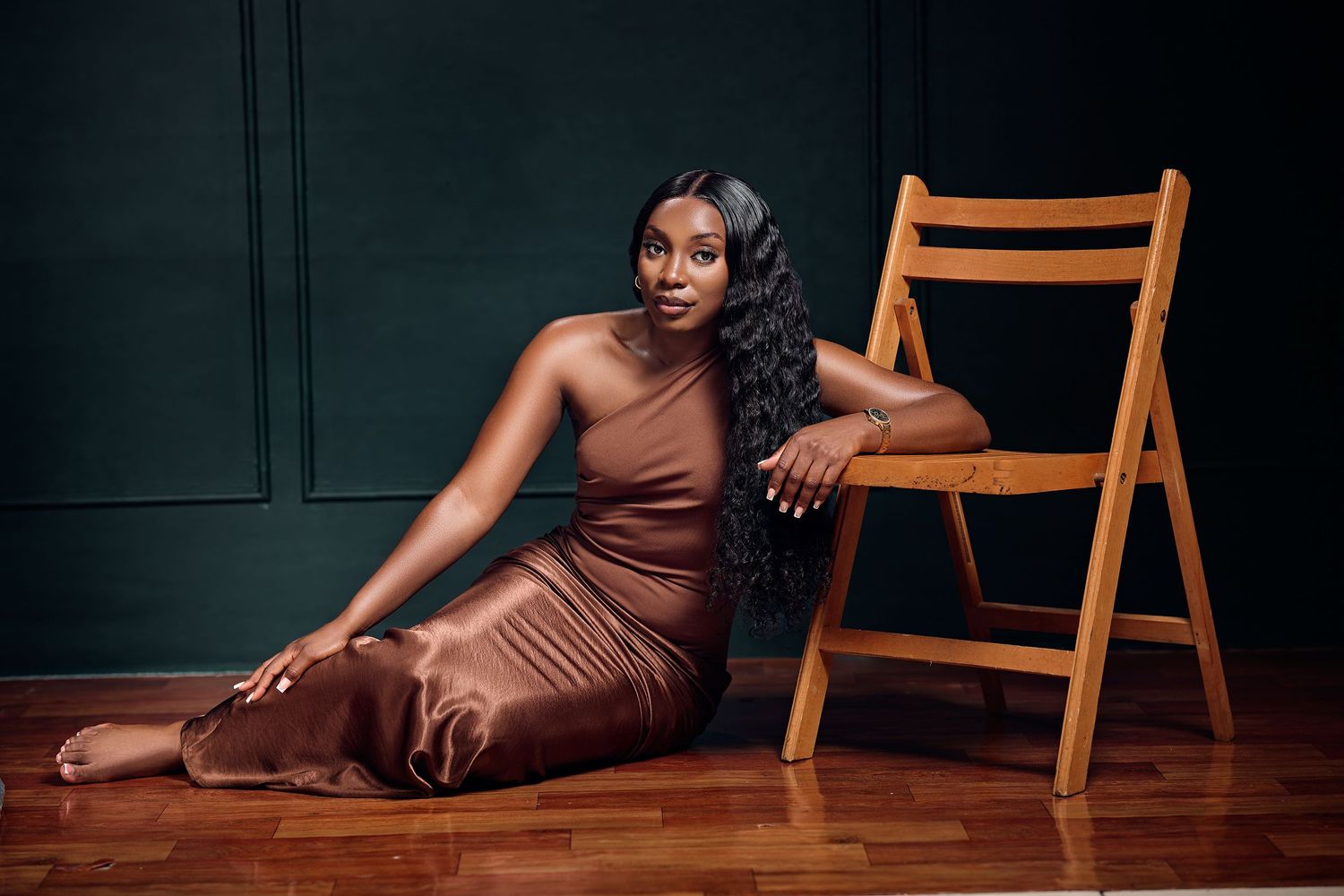Woman in brown satin outfit poses elegantly on hardwood floor beside wooden chair against dark teal wall.