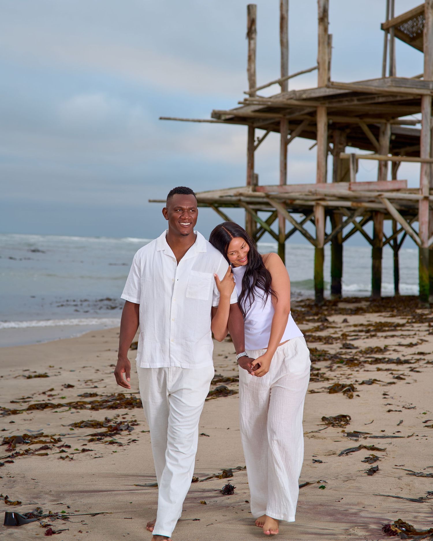 Couple dressed in white walking on a sandy beach near a rustic wooden pier with ocean waves in the background.
