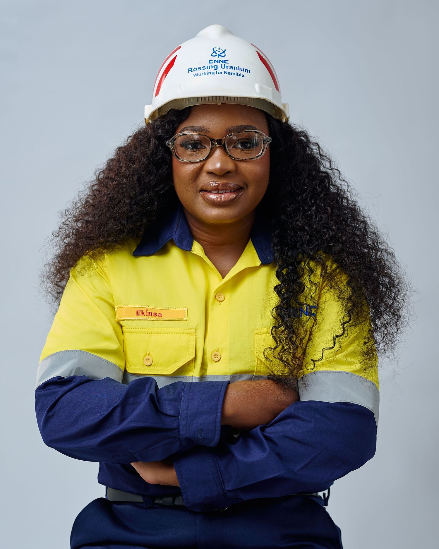 Woman in yellow and navy safety coveralls and white hard hat stands confidently with arms crossed in professional portrait.