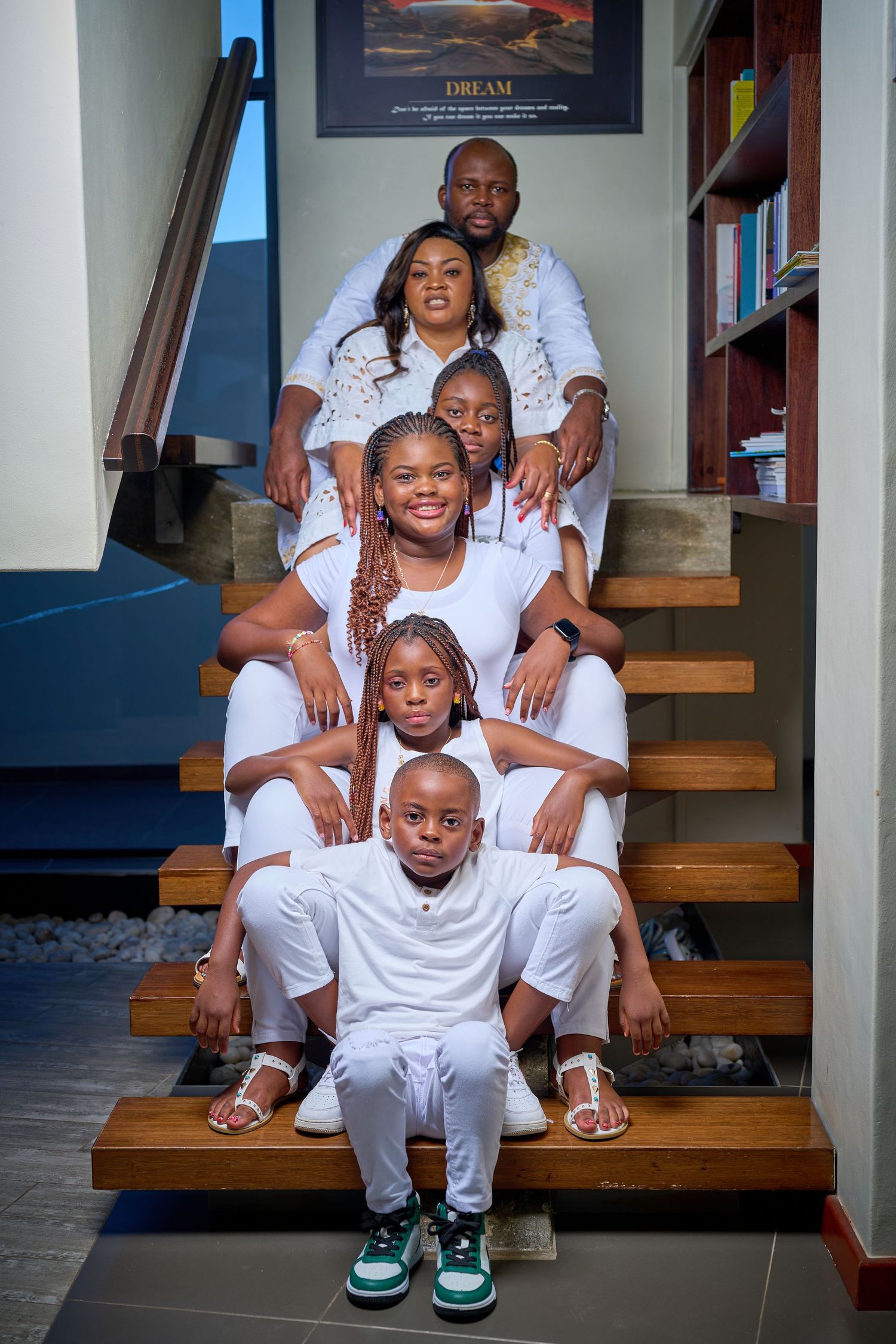 A joyful family of six poses on wooden stairs, all wearing coordinated white outfits in a modern home interior.