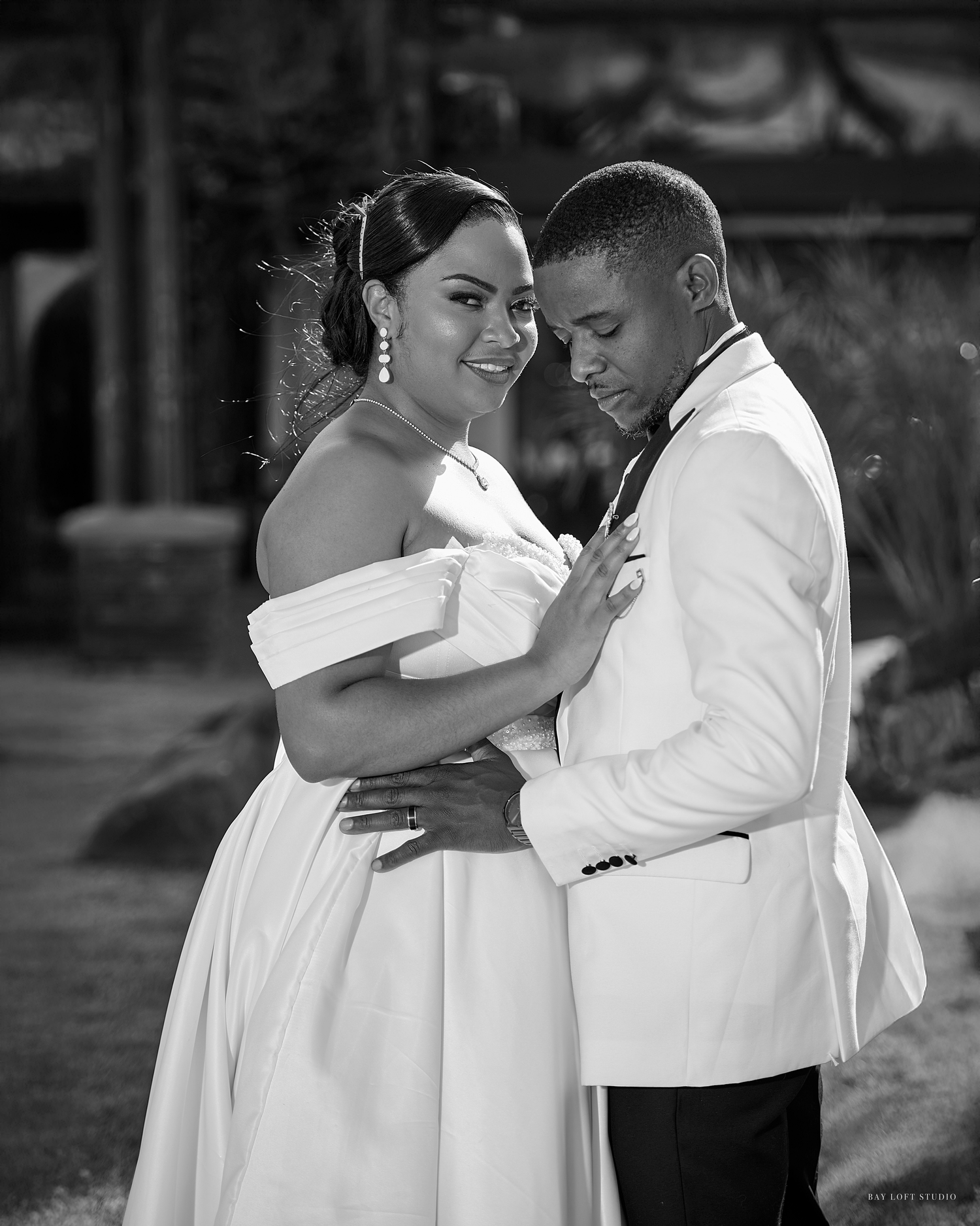 Black and white wedding photo of a couple in formal attire embracing, bride in off-shoulder gown smiling at camera.