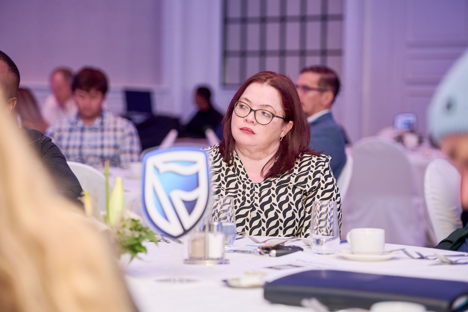 Woman with red hair and glasses attending a Standard Bank corporate luncheon event, seated at a formal table.