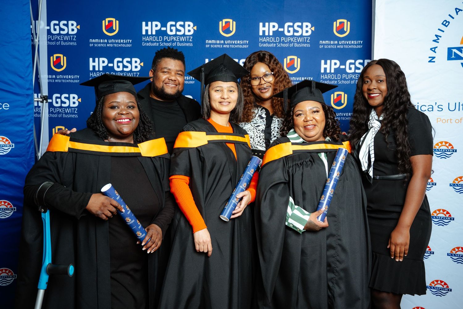 HP-GSB graduates in black and gold academic regalia celebrate together at a Namibia University graduation ceremony.