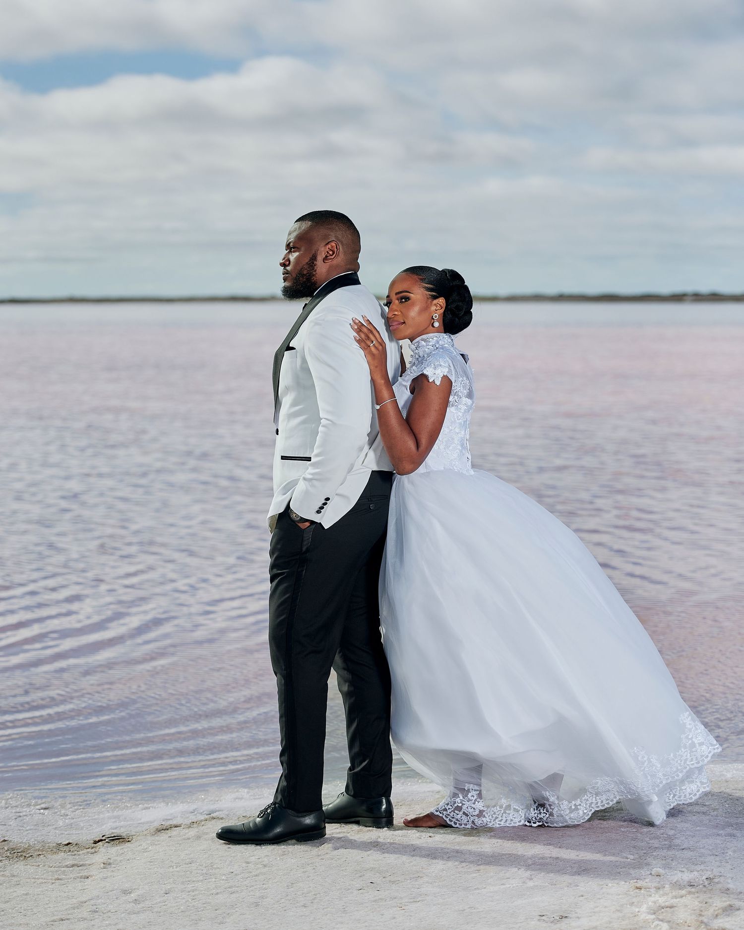 Elegant bride and groom posing back-to-back in wedding attire beside a stunning pink lake under a cloudy sky.