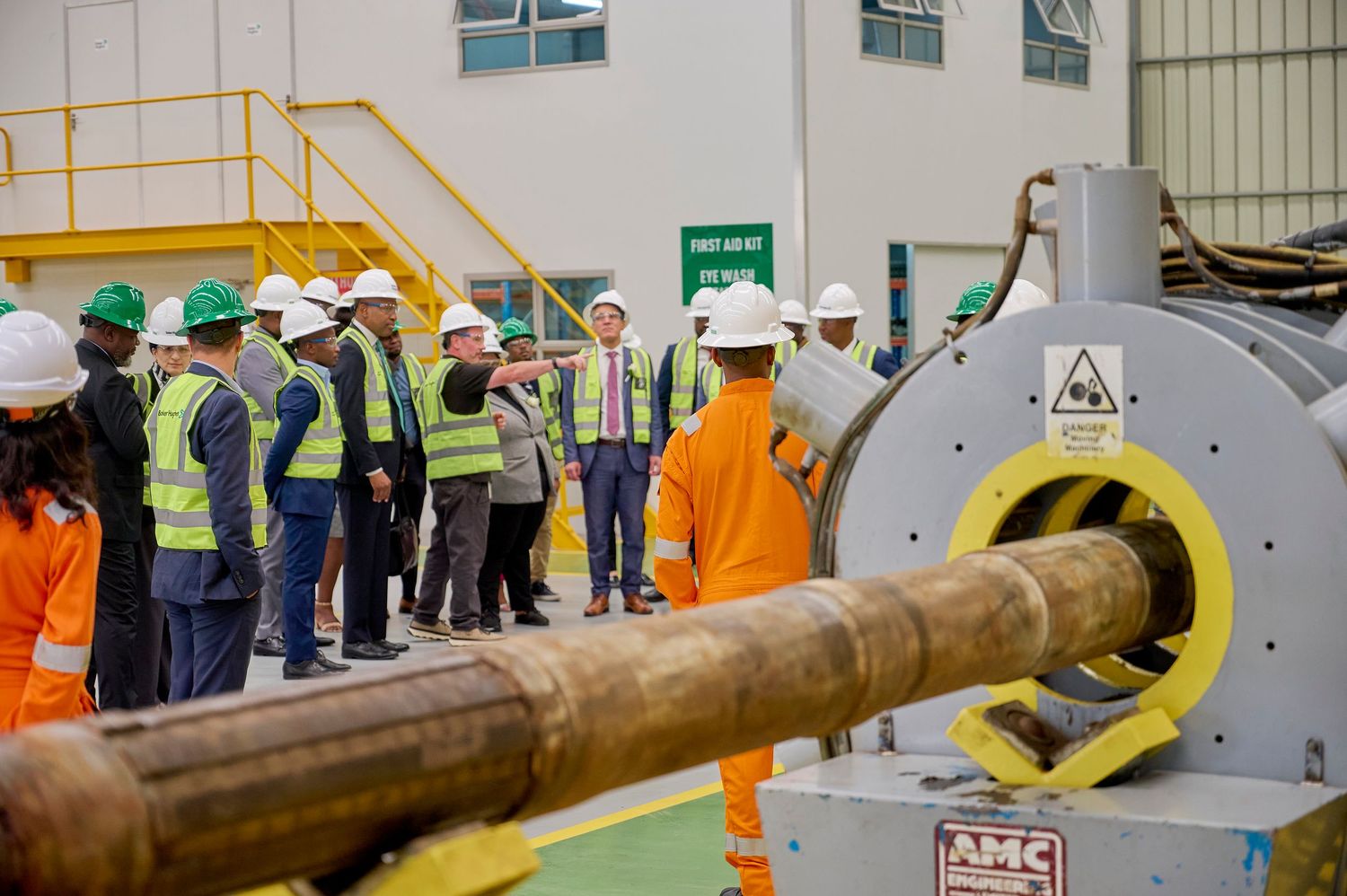 Group of workers in hard hats and safety vests touring an industrial facility with large machinery and pipes.