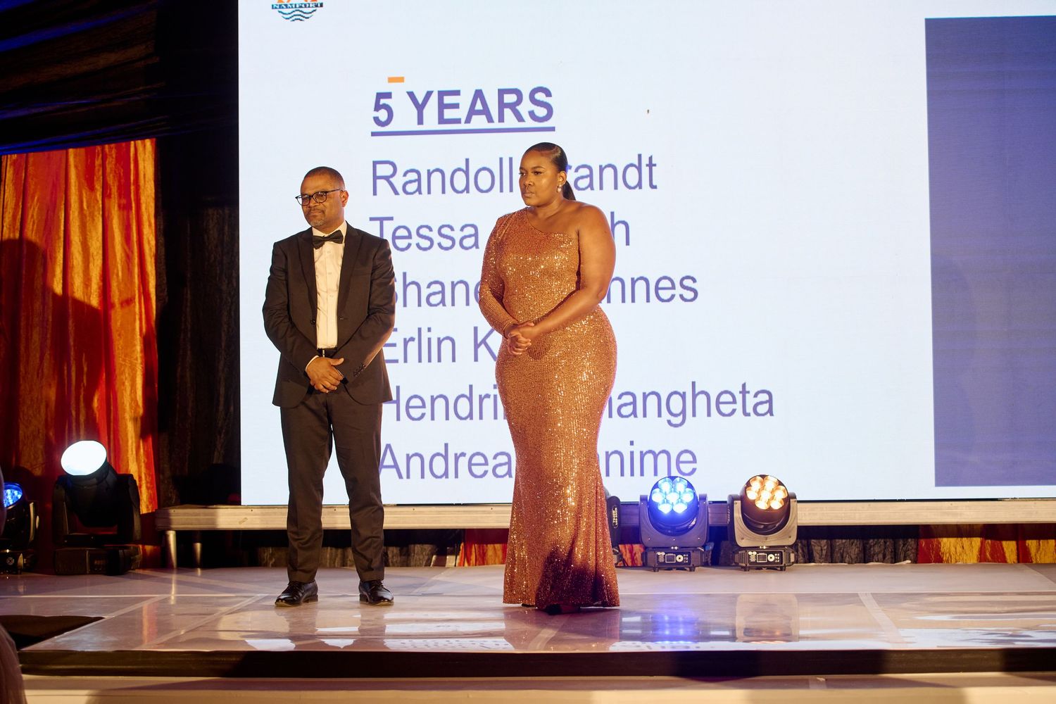 Two professionals on stage at a formal awards ceremony displaying a 5 Years employee recognition milestone list.