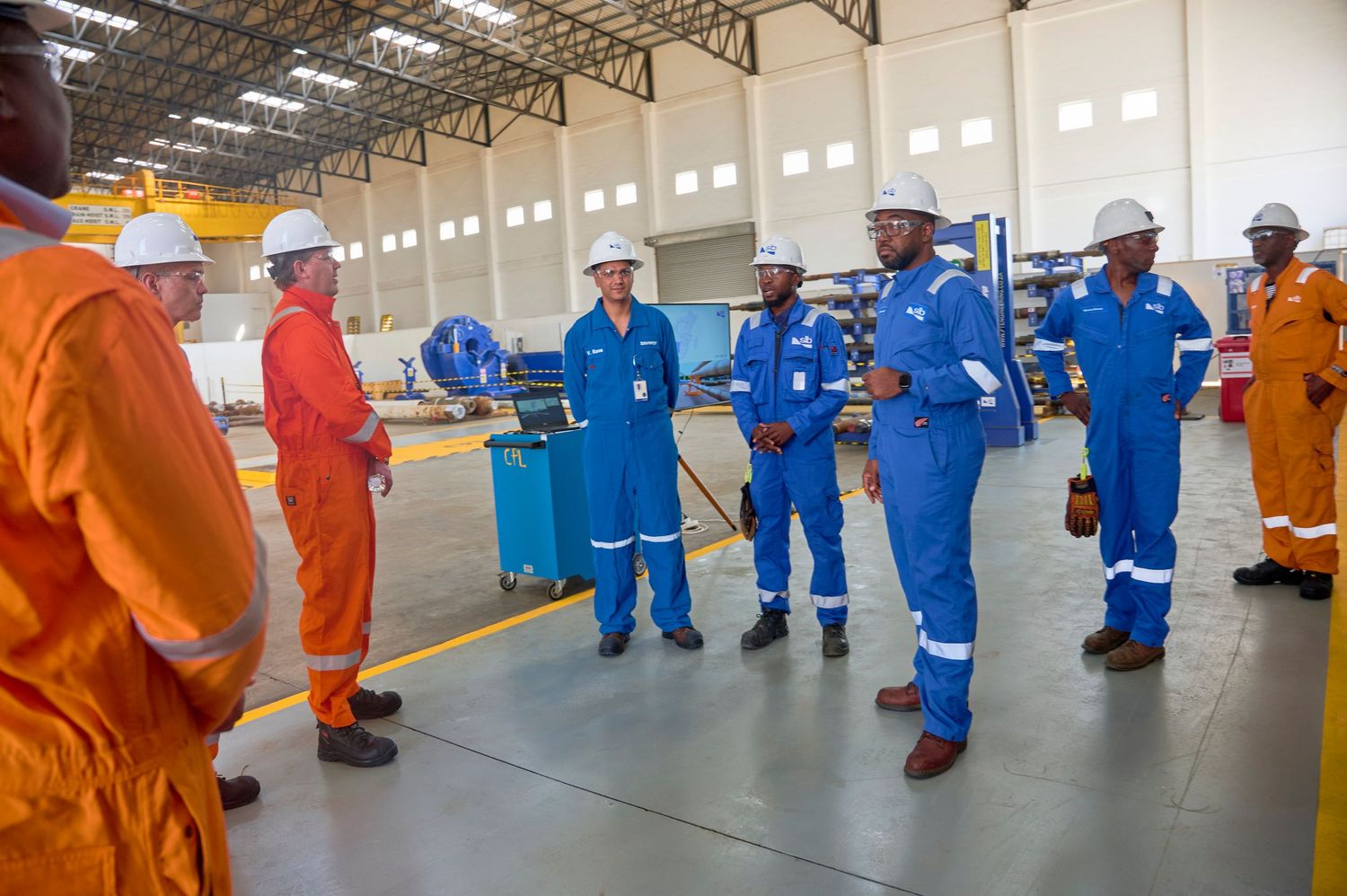 Industrial workers in blue and orange coveralls and hard hats gather for a meeting inside a large manufacturing facility.
