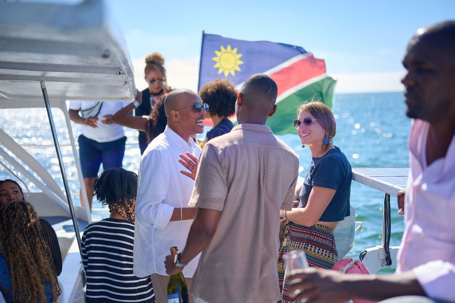 Group of people socializing on a boat with Namibia flag waving in sunny ocean backdrop.
