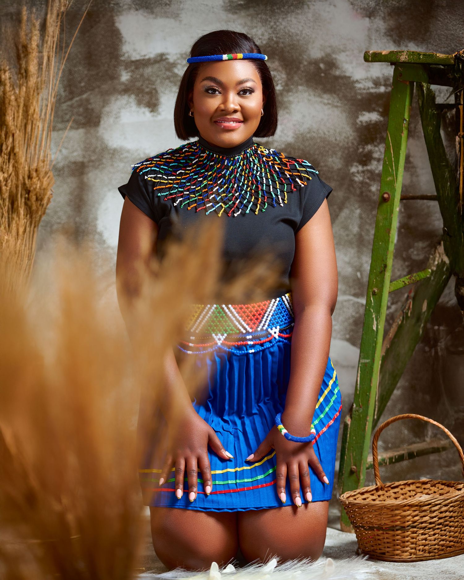A person wearing a blue and colorful beaded outfit sits in a traditional setting with a woven basket nearby.