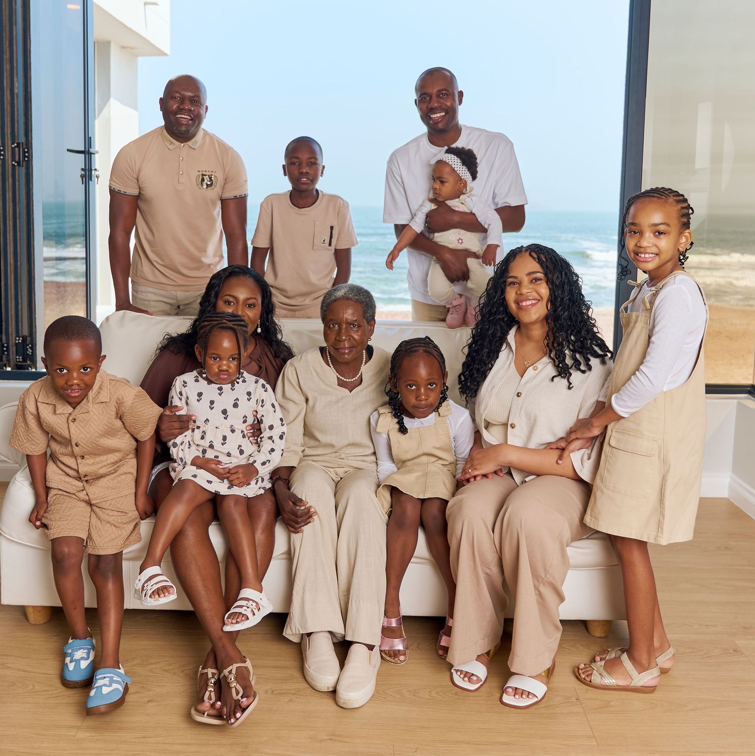 Large multigenerational family posing together indoors with ocean view, wearing coordinated neutral outfits for a portrait.