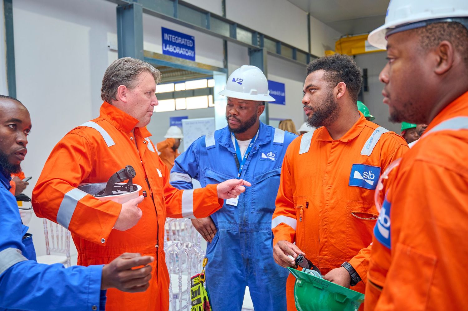 Industrial workers in orange and blue safety coveralls having a discussion inside a manufacturing facility.