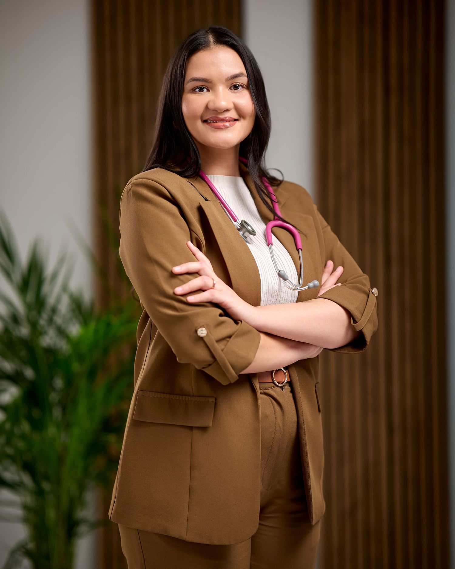 Young female doctor in brown suit with pink stethoscope stands confidently with arms crossed in modern medical office.
