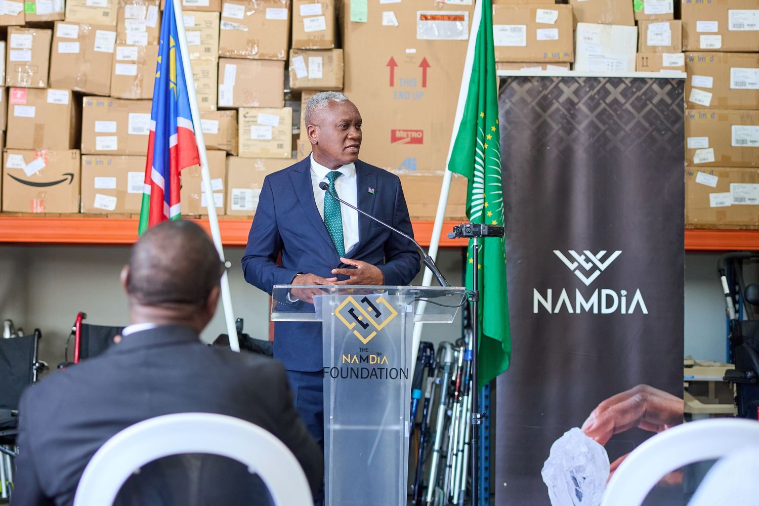 A man in a suit speaks at a podium during a NAMDIA event in a warehouse with boxes and flags.